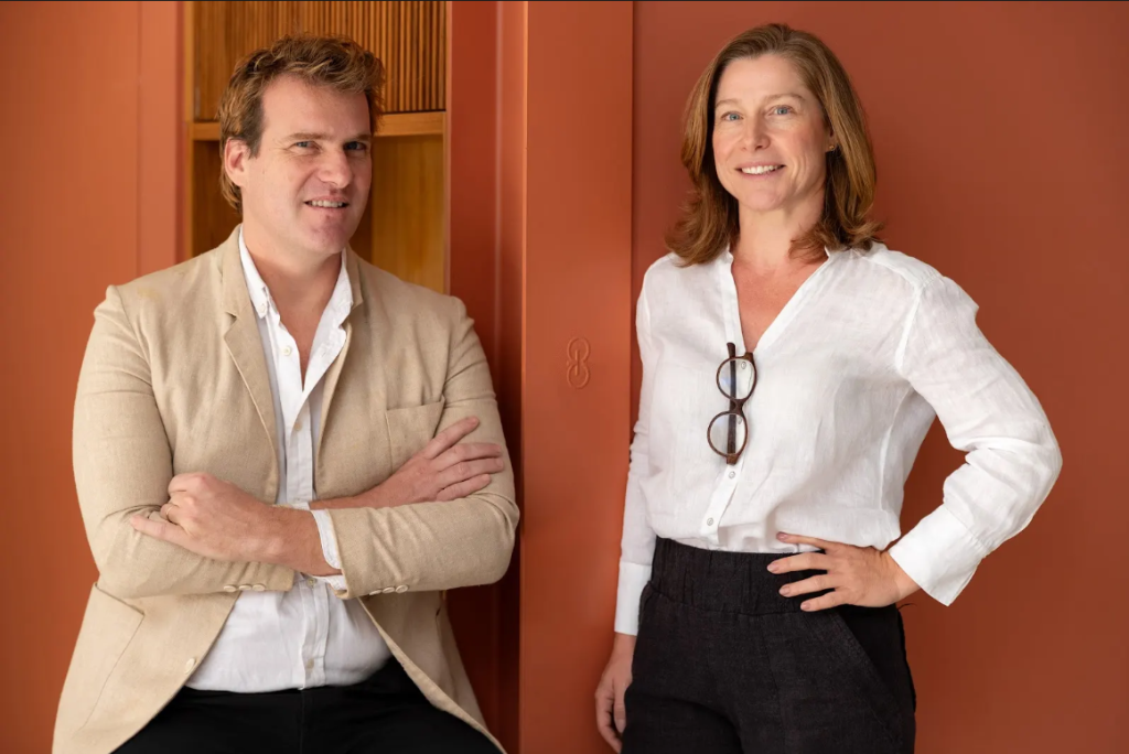 A imagem mostra duas pessoas, um homem, loiro e de olhos claros, e uma mulher, também loira e de olhos claros.
O homem está vestindo um smoking bege e a mulher está vestindo uma camiseta branca. Ambos estão em um fundo laranja.
